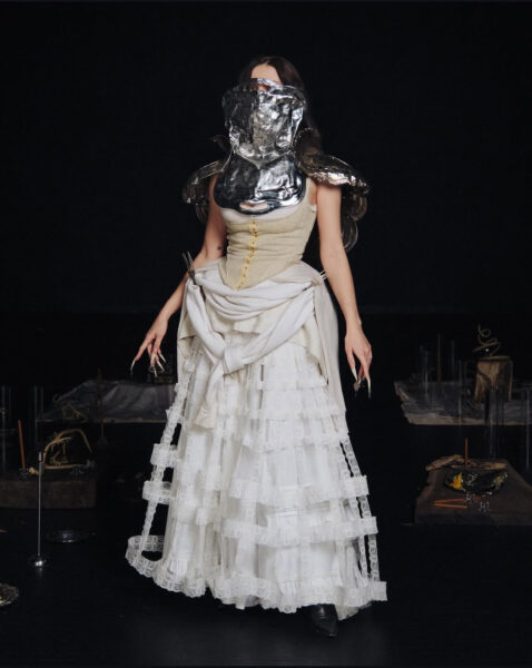 Runway look from Zoe Gustavia Anna Whalen featuring a lace gown layered with metallic armor and sculptural accessories in a dark setting.
