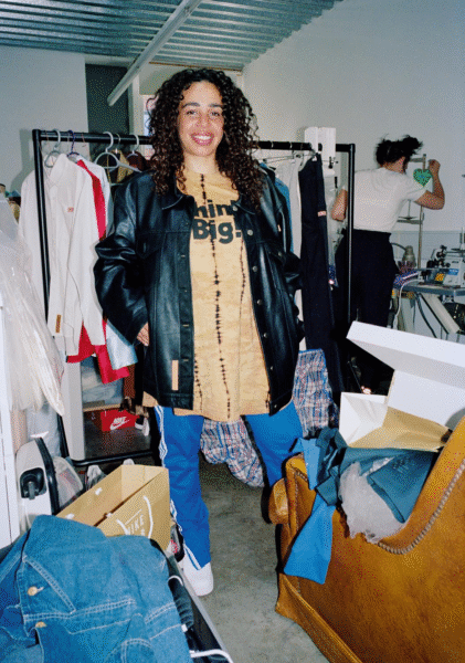 Martine Rose smiling in her London design studio, wearing a leather jacket and tie-dye shirt, surrounded by clothing racks and fashion materials. Photo by Andrea Spotorno.”