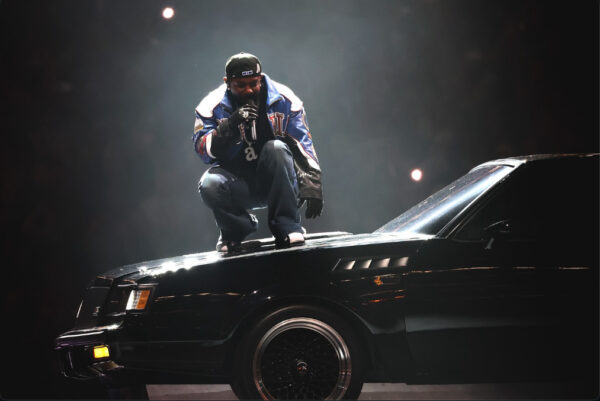 A Performer crouches on top of a black vintage car under dramatic stage lighting, wearing a bold Martine Rose jacket. Image via Martine Rose Instagram.