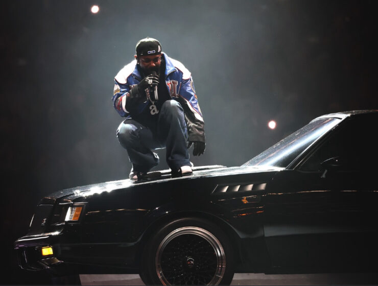 A Performer crouches on top of a black vintage car under dramatic stage lighting, wearing a bold Martine Rose jacket. Image via Martine Rose Instagram.