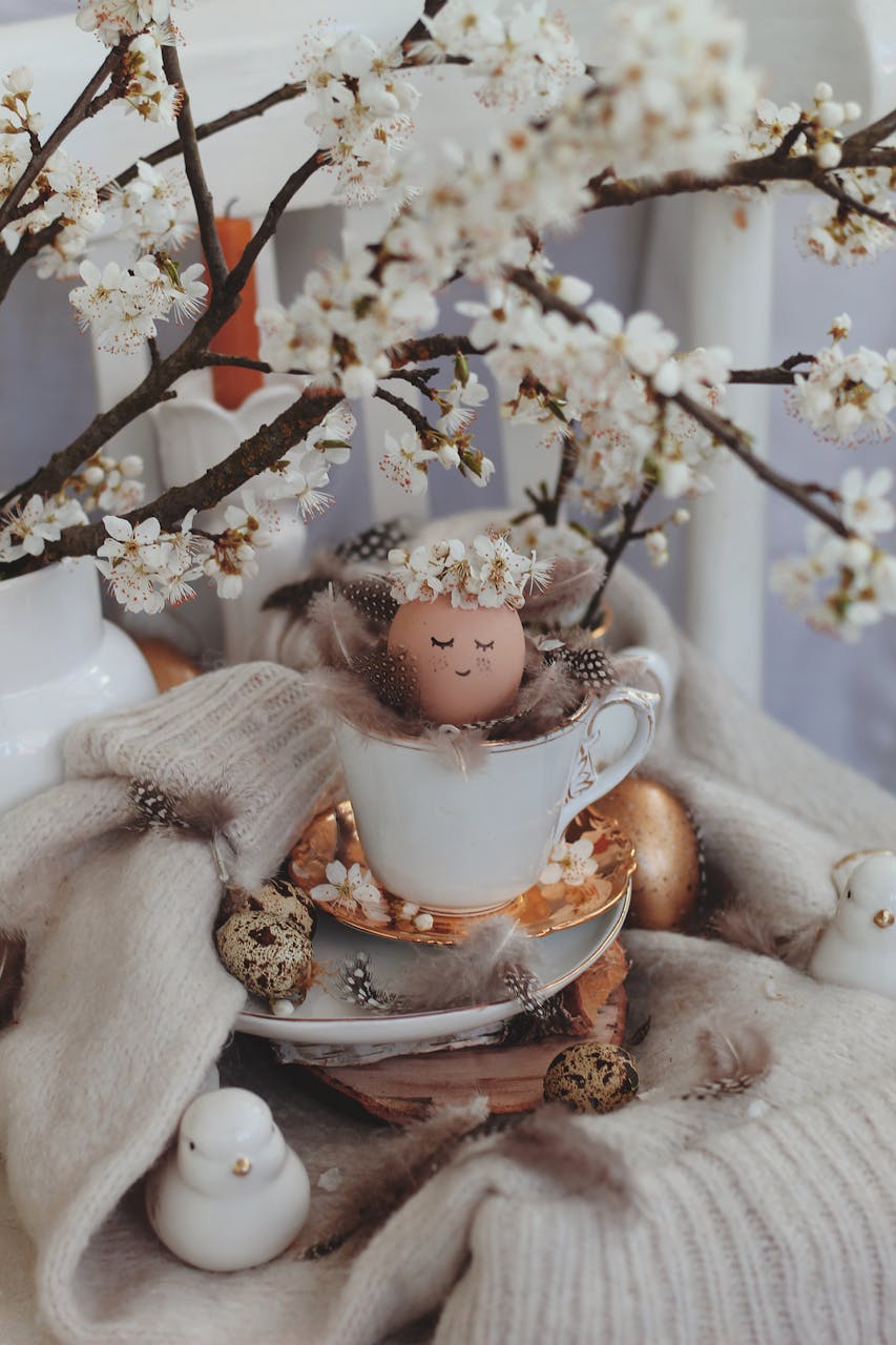 A whimsical scene April 2026 Horoscopesfeaturing a painted Easter egg in a teacup surrounded by spring blossoms and soft textures.