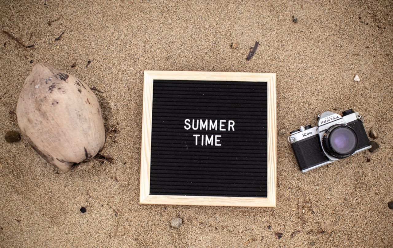 July 2026 Monthly Horoscope: Letter board with SUMMER TIME text, vintage camera, and coconut on sandy beach.
